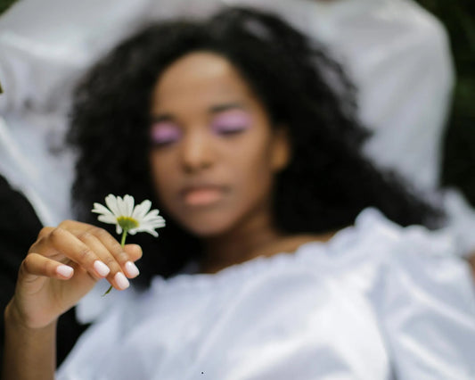 Woman laying on soft white background. She has brown skin and curly black hair, she has a pretty pink eyeshadow on her closed eyelids. She looks peaceful and about to drift off to sleep while holding a white daisy in her right perfectly manicured hand. 
