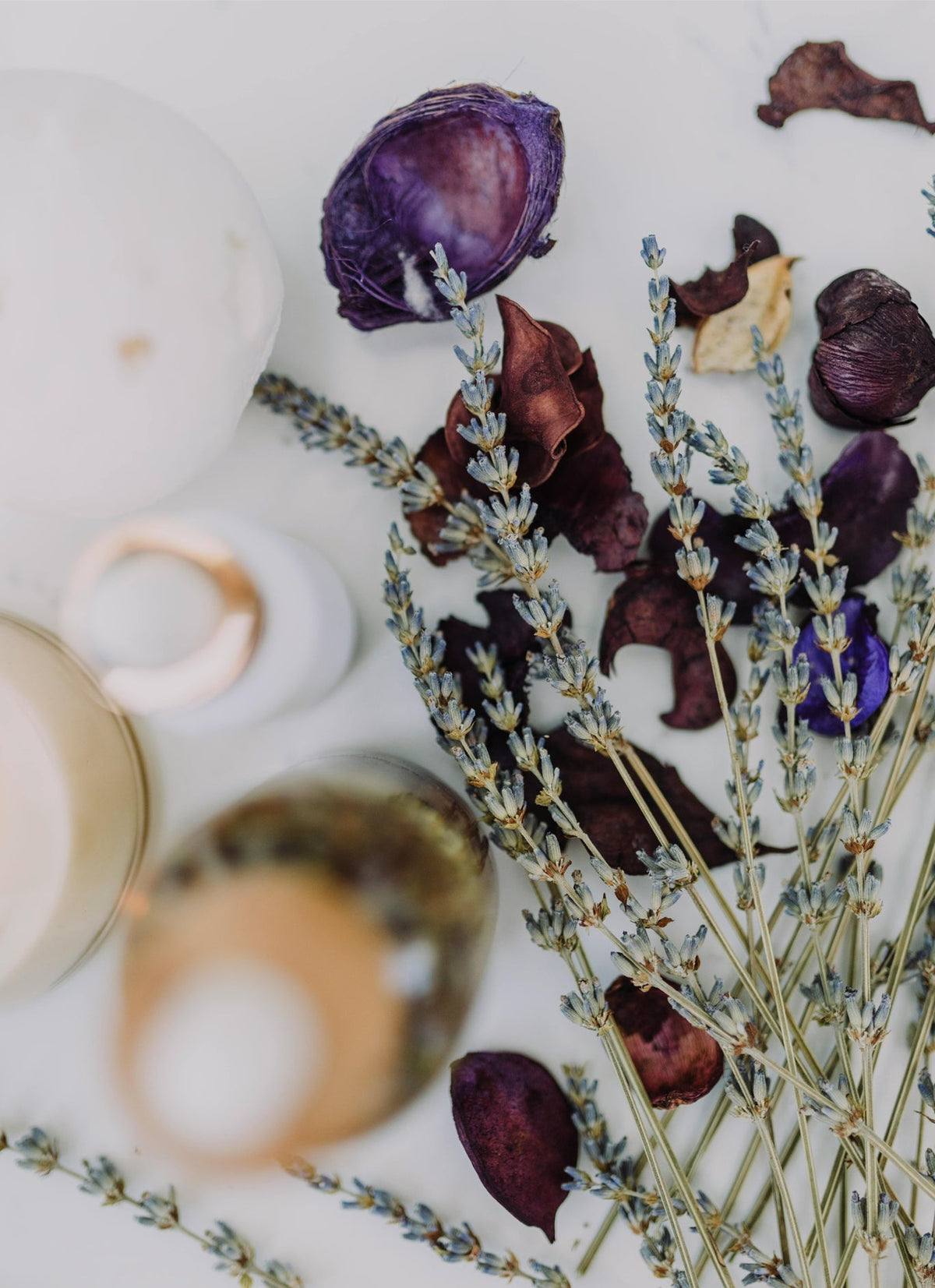 Abstract photo of dried lavender in a loos bunch is in the foreground while dark purple petals of another dried flower lay in the background alongside some candles all on a soothing white background