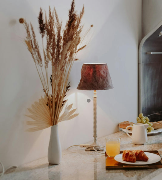 Abstract photo of a lamp and dried flower arrangement against a neutral beige wall, there is a peek of a stove and grapes on the right side of the photo