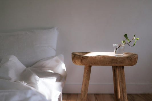 Abstract photo where the left is a bed with white pillows and linens, there is a soft rounded wooden table to the right of the bed, with a clear glass holding a small plant cutting. Sunlight strikes both the table and the bed in a diagonal dreamy manner