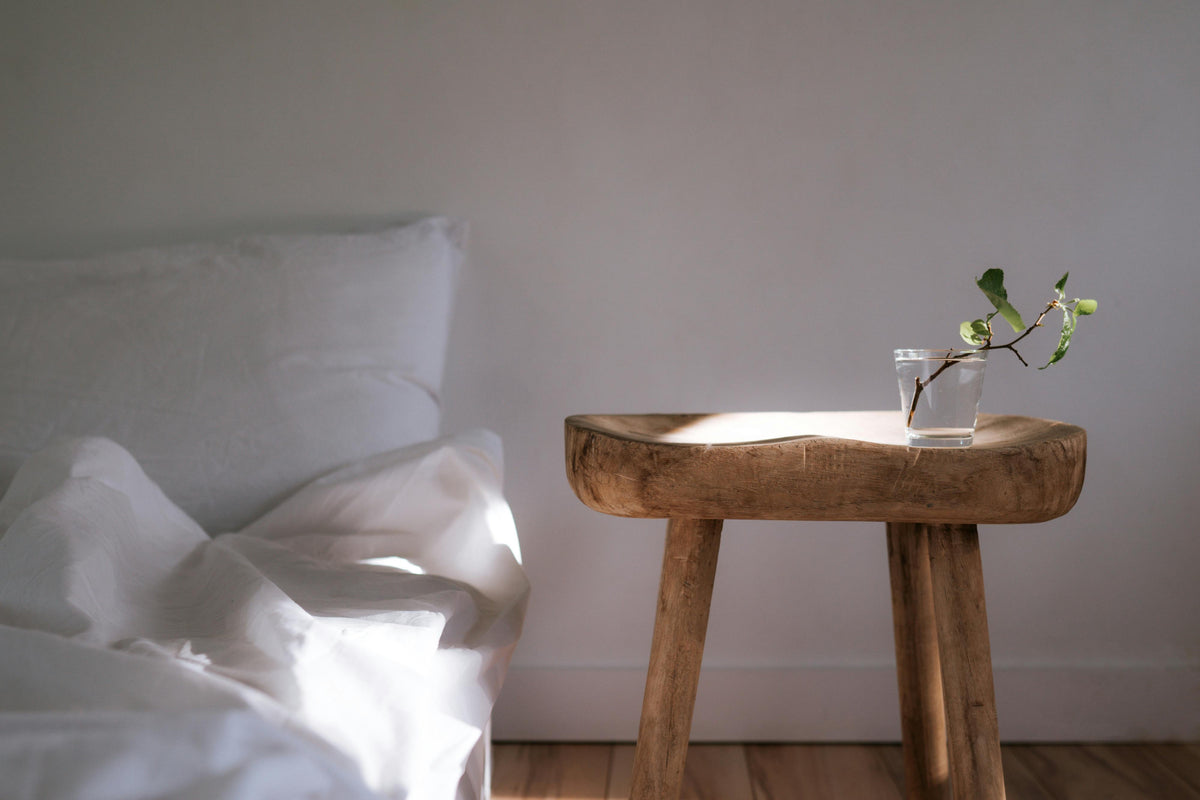 Abstract photo where the left is a bed with white pillows and linens, there is a soft rounded wooden table to the right of the bed, with a clear glass holding a small plant cutting. Sunlight strikes both the table and the bed in a diagonal dreamy manner