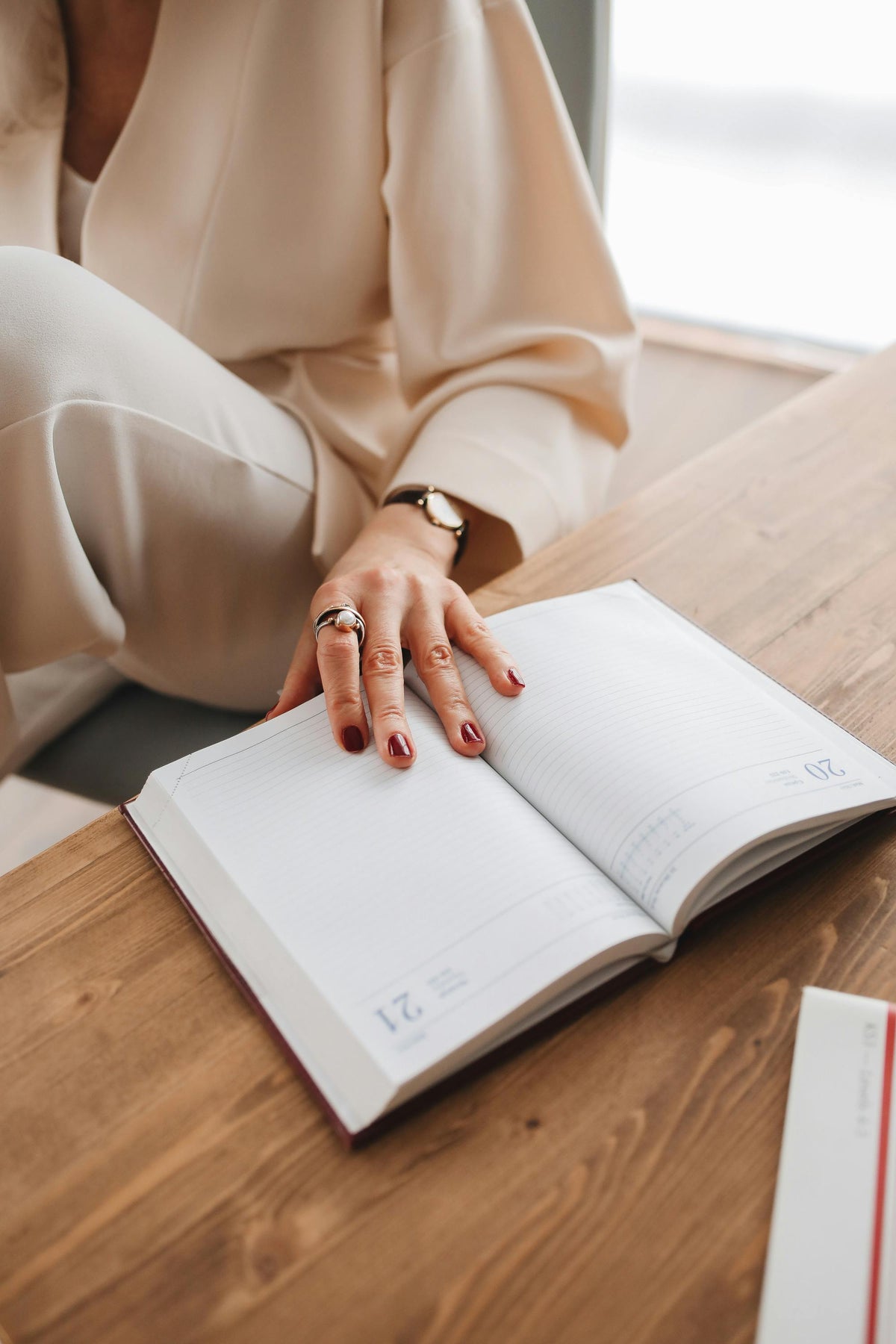 Person with their manicured hand (painted dark red finger nail polish), on a white lined journal page, on a wooden desk and the person is wearing neutral tones. The hand has a ring on the index finger and a simple watch on the wrist.